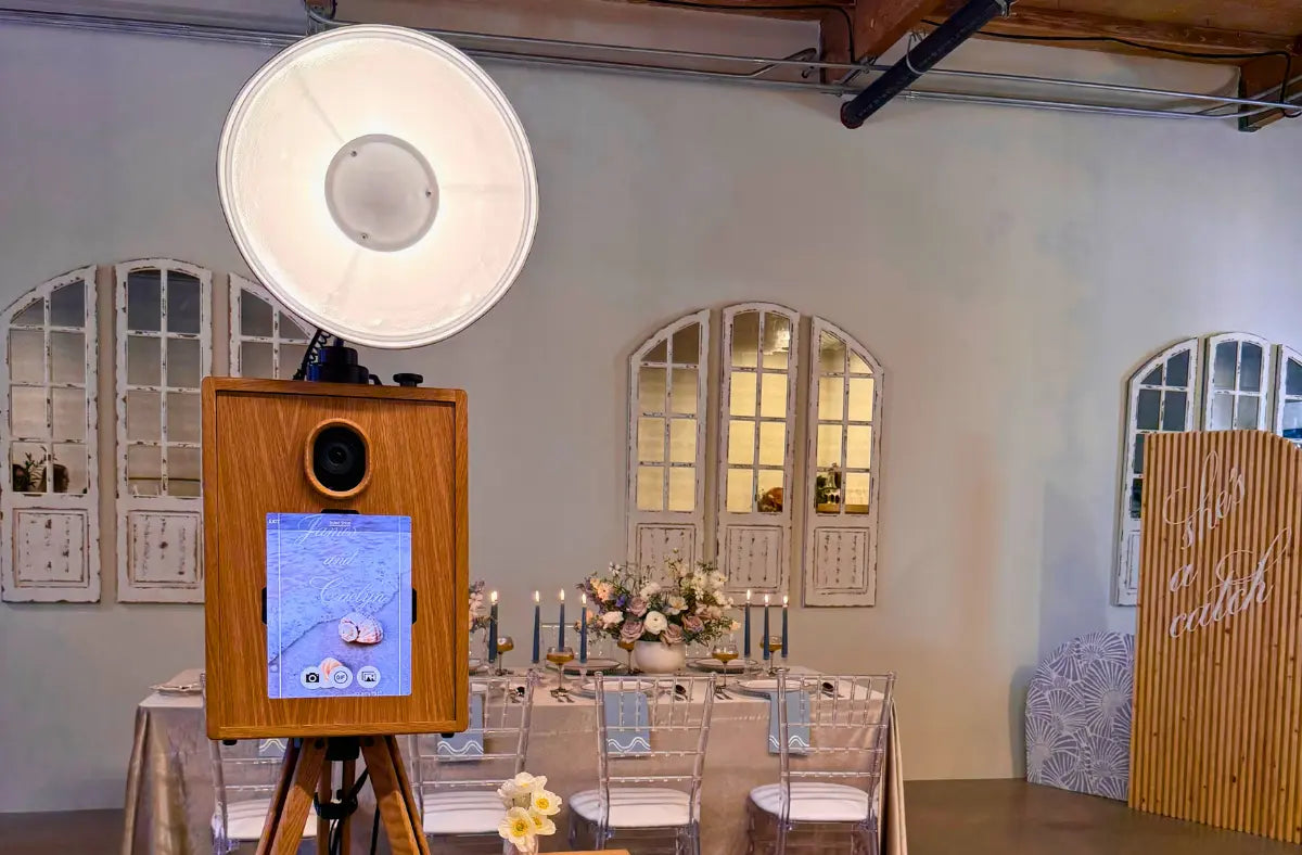 A wooden photo booth with a large ring light sits in front of a decorated dining table with chairs. There is a bamboo partition with text on the right side of the room.