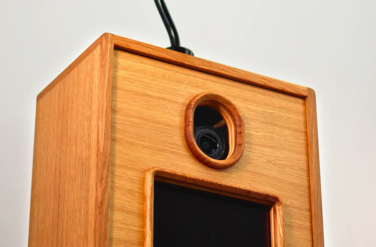 A close-up of a wooden photo booth with a camera lens visible through a circular opening and a dark screen below.