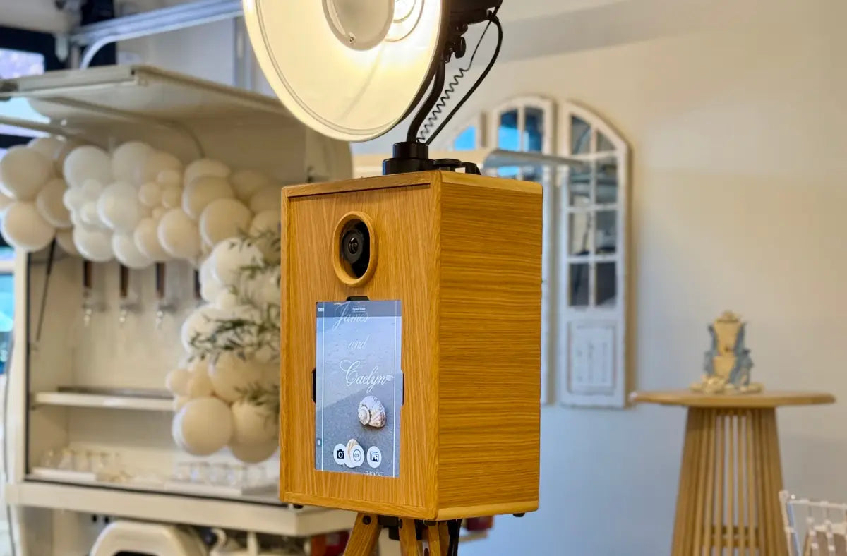 A wooden photo booth with a touchscreen and camera lens is set up near a bar decorated with white balloons and greenery.