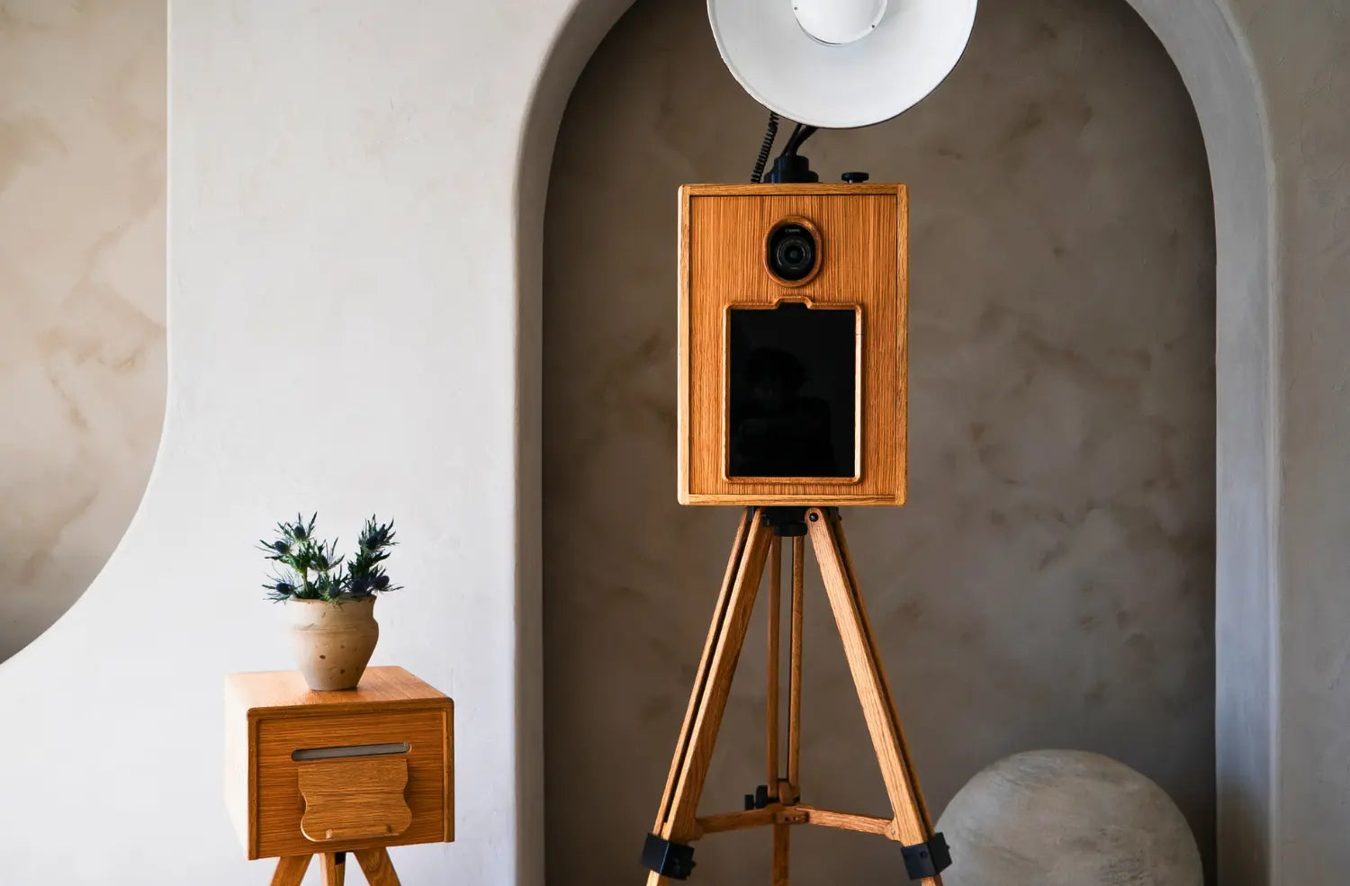 A wooden glam photo booth on a tripod, with a ring light above it and a wooden printer frame beside it, set against a textured wall.